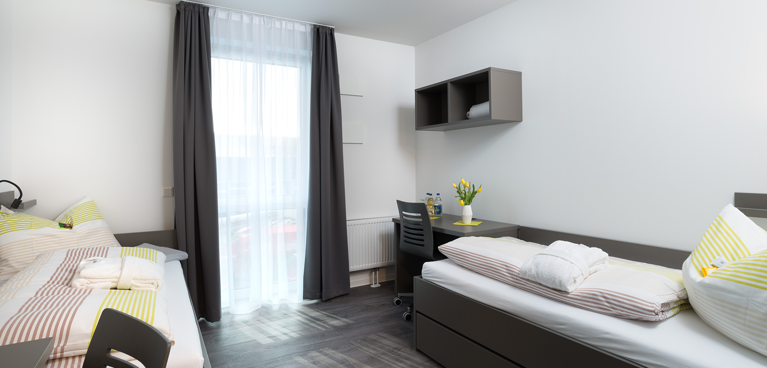 Ein modernes, aufgeräumtes Zweibettzimmer mit zwei Einzelbetten, gestreifter Bettwäsche, einem Schreibtisch mit Stuhl, Regalen, einem Fenster mit Verdunklungsvorhängen und einer Vase mit gelben Tulpen auf dem Schreibtisch.