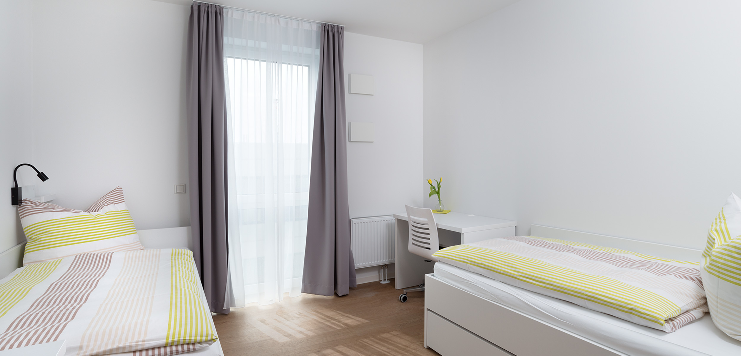 Ein helles, minimalistisches Schlafzimmer mit zwei Einzelbetten, jedes mit gestreifter Bettwäsche in Gelb, Weiß und Beige. Ein Schreibtisch mit einem weißen Stuhl und einer Vase mit Blumen steht am Fenster, das mit durchsichtigen Vorhängen und grauen Gard